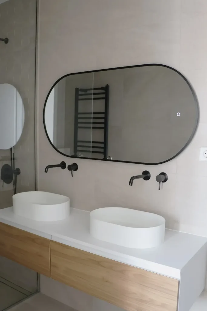 Baño con doble lavabo sobre mueble de madera clara, grifería negra y espejo ovalado con marco negro