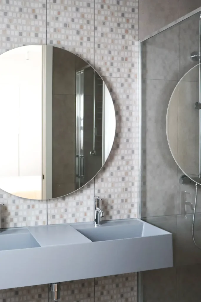 Baño con lavabo suspendido en gris azulado, espejo redondo, pared de mosaico y mampara de ducha de cristal