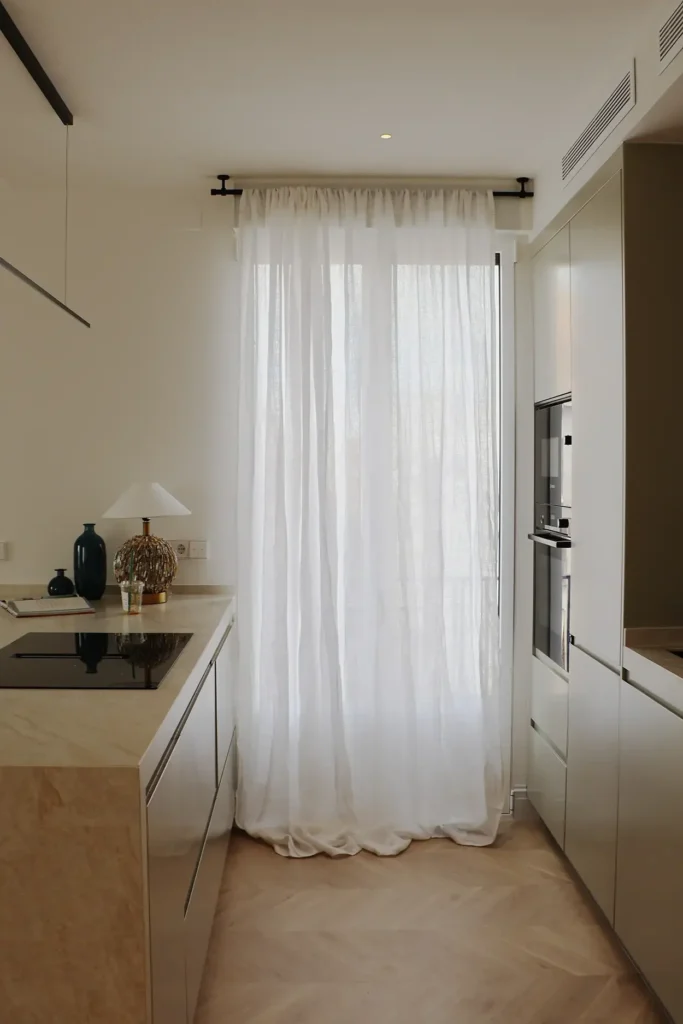 Cocina con armarios en verde salvia, placa de inducción, cortina de lino blanca y parquet en espiga