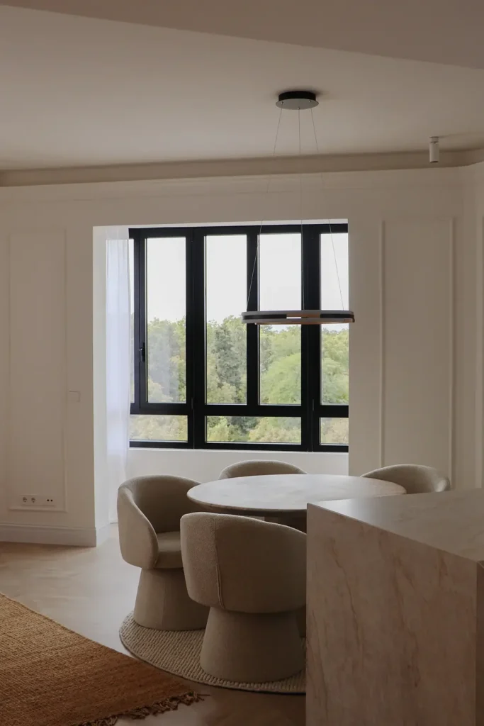 Zona de comedor con mesa redonda y sillas tapizadas junto a una ventana de carpintería negra con vistas al arbolado