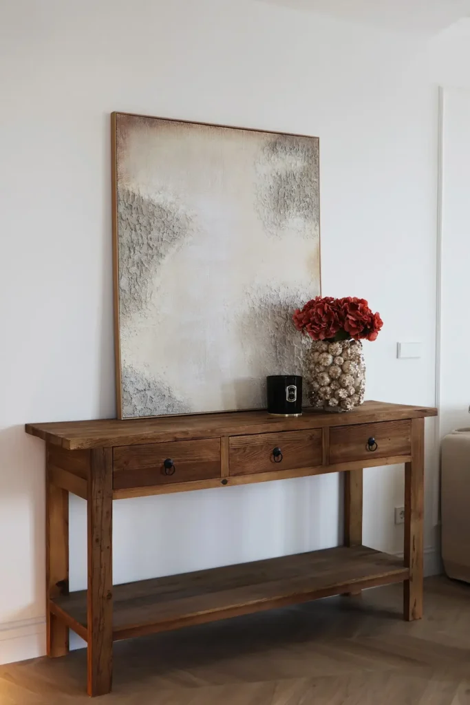 Consola de madera envejecida con cuadro abstracto y jarrón con flores rojas sobre suelo de parquet en espiga