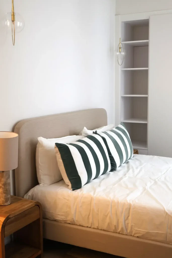 Dormitorio con cama de cabecero tapizado beige, cojines a rayas verdes, mesita de madera y estantería empotrada