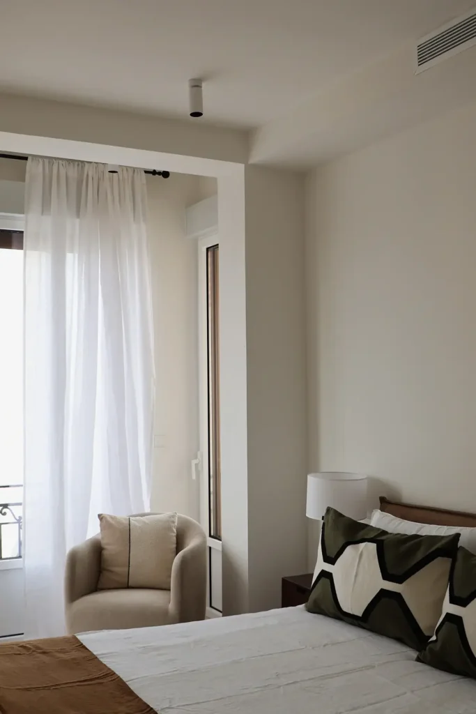 Dormitorio con cama de ropa blanca y cojines estampados, butaca tapizada y cortinas de lino junto a la ventana