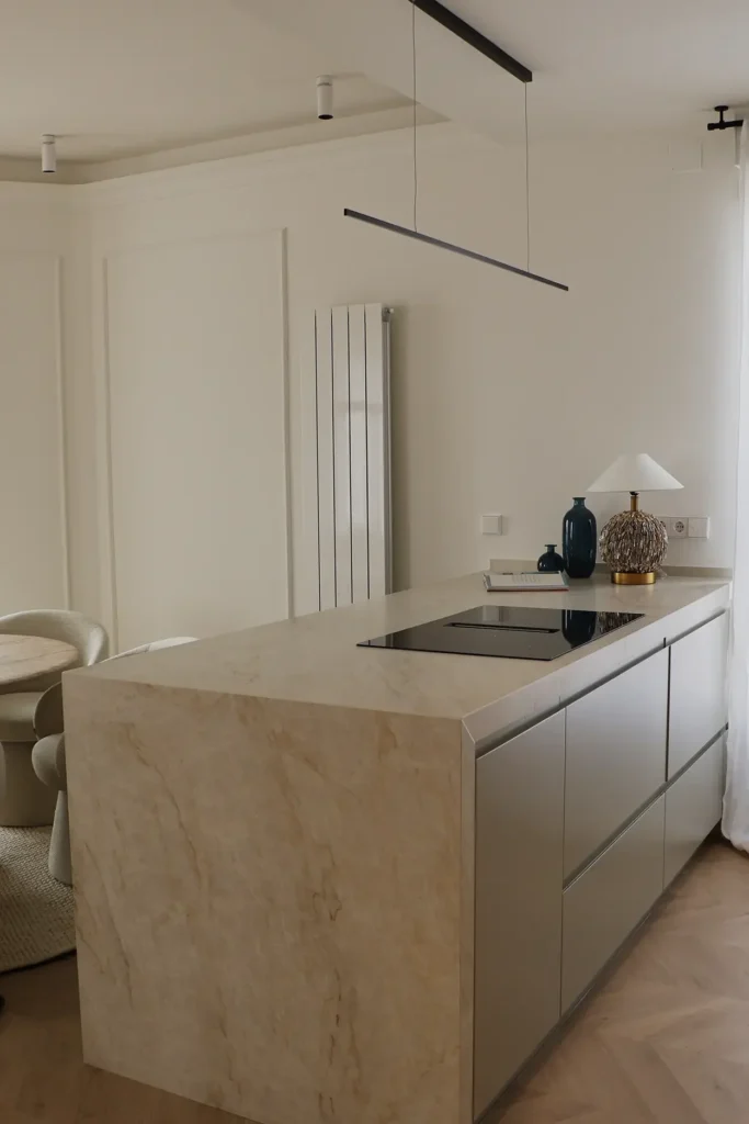 Isla de cocina con placa de inducción, frentes en verde salvia, lateral de piedra veteada y lámpara lineal negra