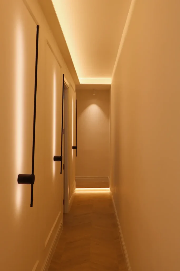 Pasillo con iluminación indirecta cálida, apliques lineales negros y suelo de parquet en espiga
