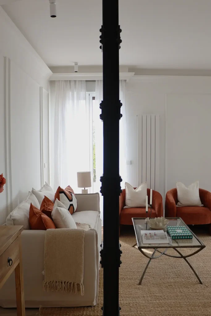 Vista del salón con columna metálica negra, sofá beige, butacas terracota y alfombra de fibra natural