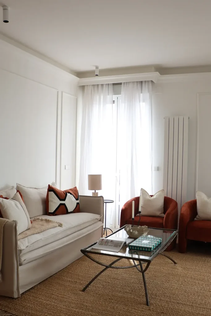 Zona de estar del salón con sofá, mesa de centro de cristal y cortinas de lino blancas junto al ventanal